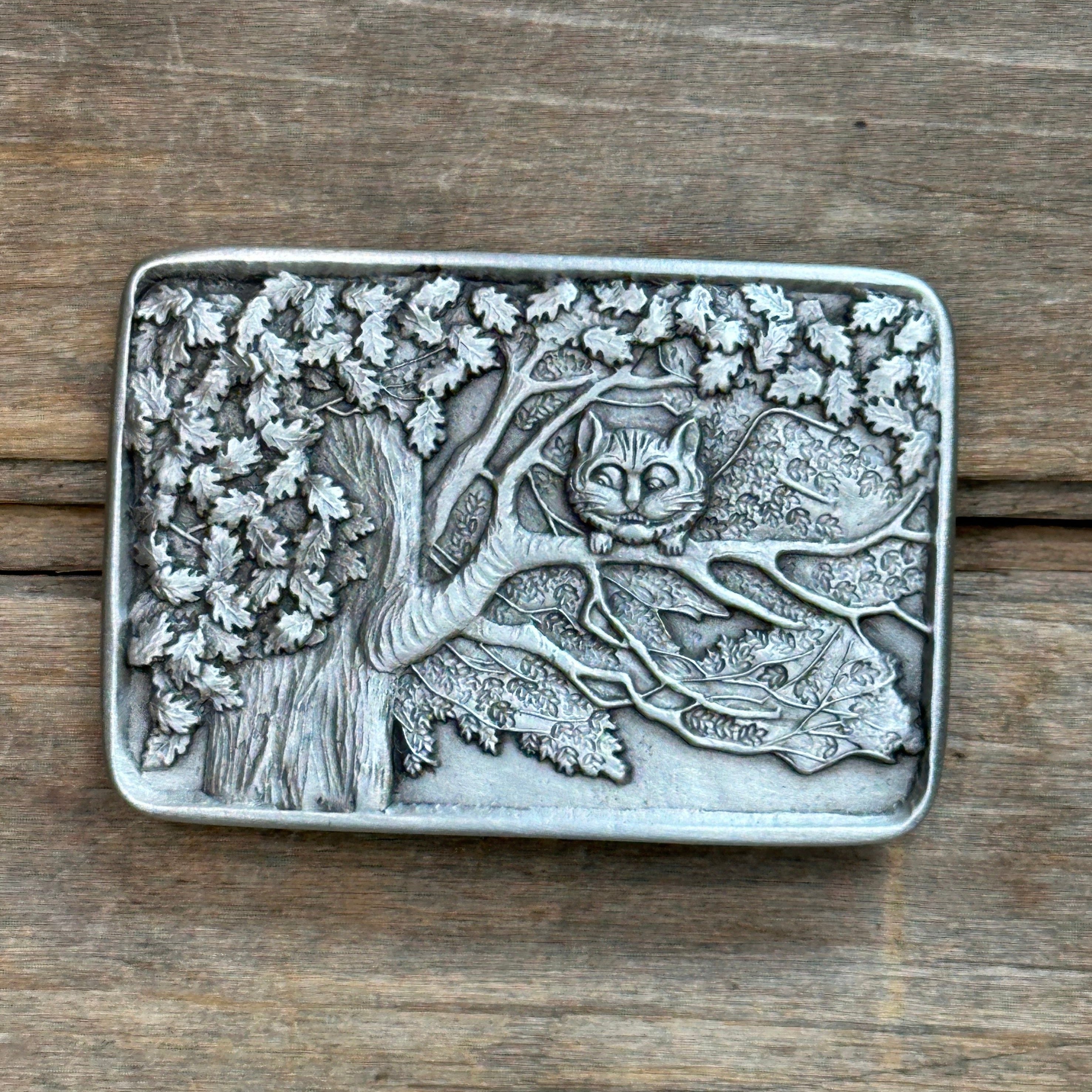 This is a pewter belt buckle with silver tone.   It Depicts the Cheshire Cat, Grinning, Perched in a tree.