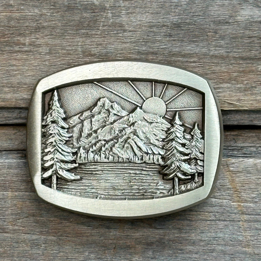 This is a solid brass belt buckle with a gold tone and brushed finish.  It depicts tall long leaf pines overlooking a lake and mountain and sunrise.