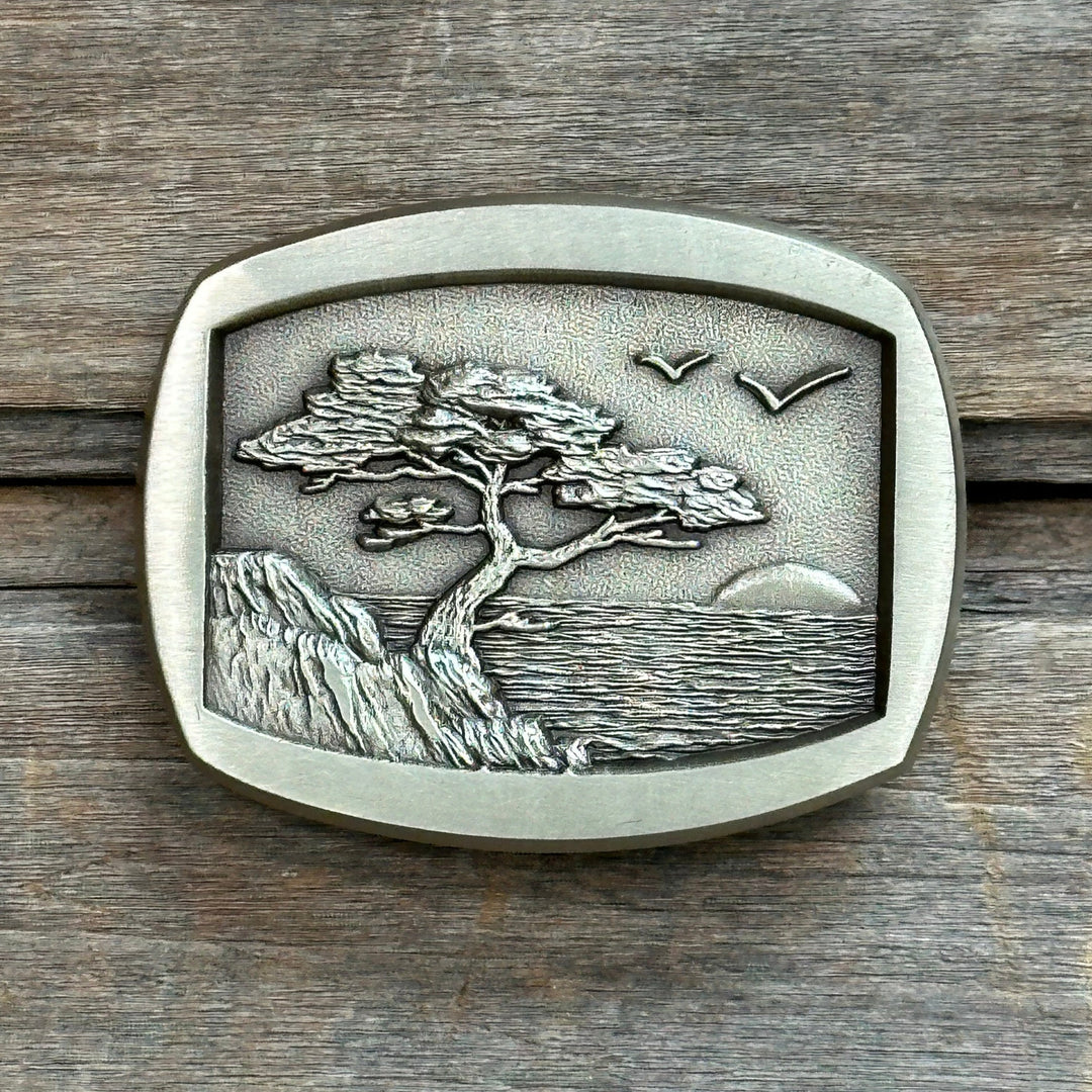 This is a solid brass belt buckle with a brushed finish and soft gold tone. It depicts a rugges western coastline with a tree clinging to a cliff over lookind the ocean and a sunset.