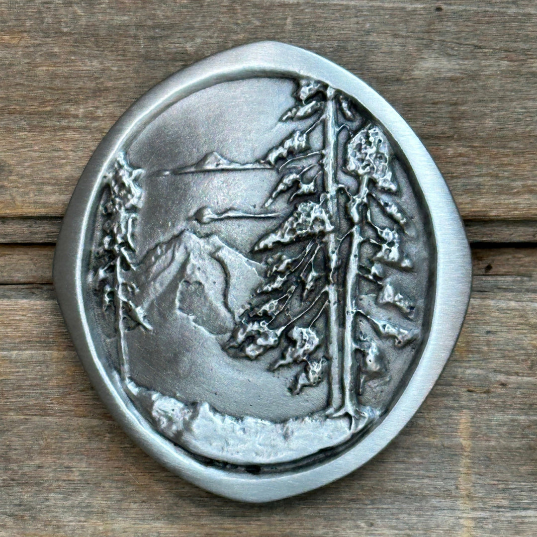 This is a pewter belt buckle with a sliver tone.  It depicts a mountian scene with tall pines in the fore ground and clouds above.