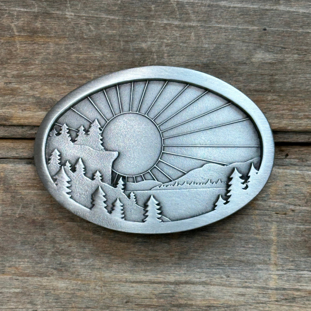 This is a pewter belt buckle with a silver tone.  It depicts a mountain scene with sunrays shining down on the mountains, trees, and a lake.